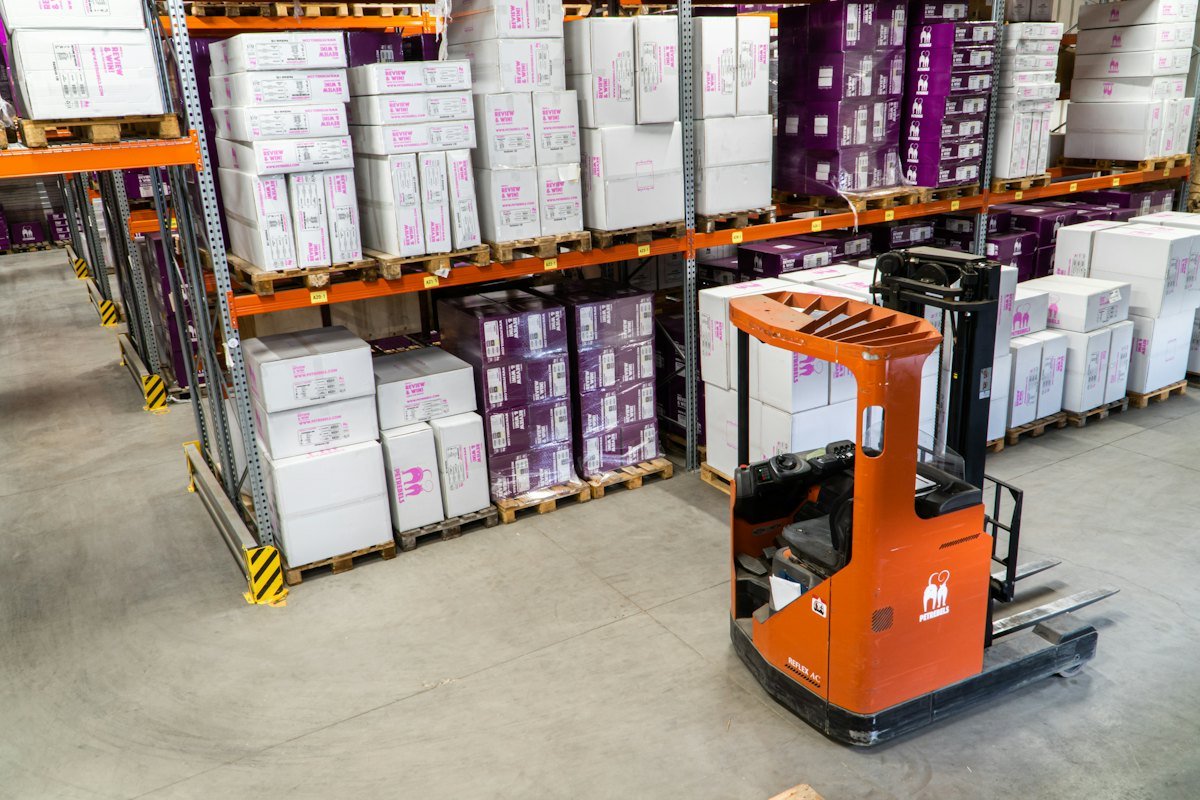 An orange forklift parked beside pallets of white and purple cartons on warehouse racking